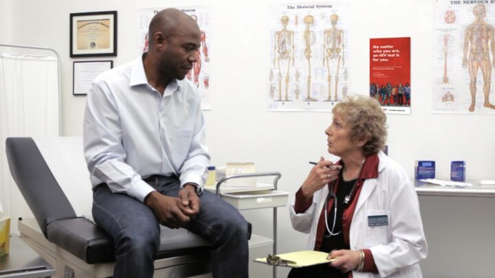 A doctor consults with a patient sitting on an examination table in a medical office, with anatomical charts on the walls, discussing the importance of regular HIV screening as part of efforts to end the HIV epidemic.