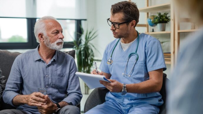 In a clinical setting, an HCP in blue scrubs talks with an older patient holding a glass of water, addressing concerns amid the Adderall shortage.