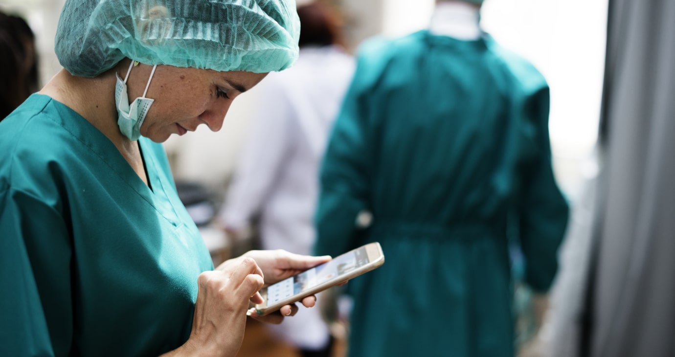 A medical professional in scrubs and a hair cover uses a smartphone, possibly utilizing AI for medical diagnosis. Two more individuals in medical attire stand in the background, contributing to this innovative approach in predicting patient outcomes.