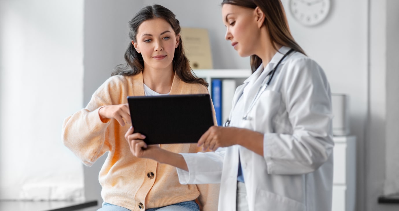 A doctor in a white coat uses AI on a tablet to show a seated woman in a light sweater, predicting patient outcomes during their consultation in the medical office.