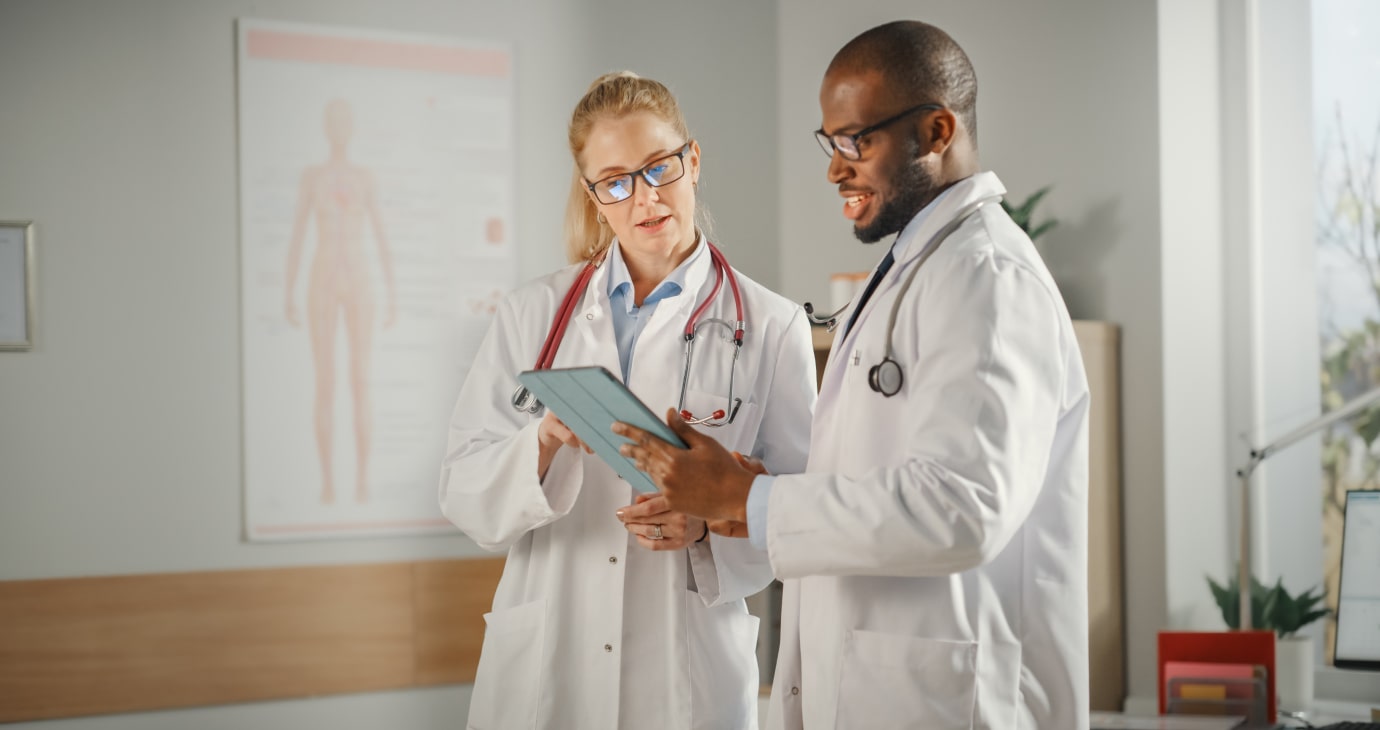 Two doctors in white coats discuss information on a tablet utilizing medical AI in their office. A poster of the human anatomy is visible in the background, underscoring how AI for medical diagnosis is revolutionizing patient care.
