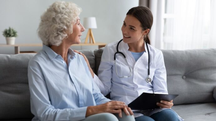 An older woman sits on a couch conversing with a female doctor, clipboard in hand. The neurologist, with a stethoscope around her neck and a reassuring smile, discusses cutting-edge Alzheimer's disease treatments.