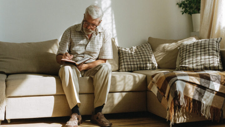 An elderly man with glasses sits on a sofa, writing in his notebook. The room is well-lit, adorned with cushions and a cozy blanket. His notes reflect the latest Alzheimer's trends, highlighting emerging amyloid-targeting antibodies and promising blood biomarkers research.