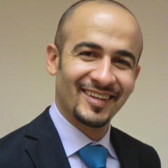 Amer Harky, FRCS, a man in a suit and tie, smiles at the camera against a plain beige background.