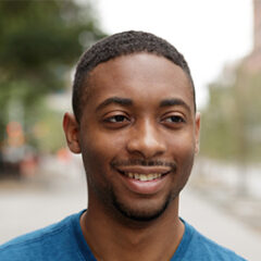 A man with short hair and a beard smiles slightly while wearing a blue shirt. He is outdoors, offering subtle business insights against a blurred backdrop of trees and buildings.
