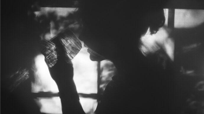 A silhouetted person rises to sip from a glass in front of a window, where dappled light and shadows dance elegantly.