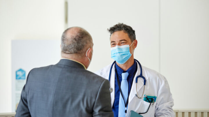 The doctor, a dedicated healthcare provider, wearing a mask and stethoscope, discusses important topics with a man in a suit indoors.