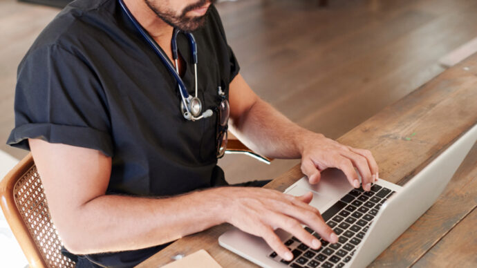 A person in medical scrubs and a stethoscope types on a laptop, diving into business insights at a wooden desk.
