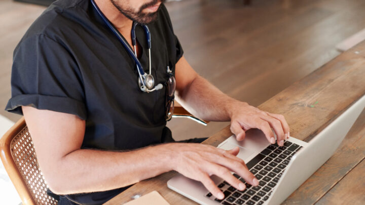 A person in medical scrubs and a stethoscope types on a laptop, diving into business insights at a wooden desk.
