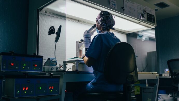 A person wearing scrubs, likely one of the dedicated physicians, is seated at a workstation, intently looking through a microscope. Amidst the various equipment and monitors surrounding the station, there's an air of diligence reminiscent of early COVID investigation days.