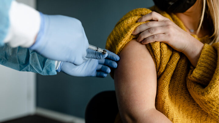 A person in a yellow sweater is receiving a COVID-19 vaccine injection in the arm from a healthcare worker wearing gloves, highlighting the potential promise of digital immunity passports for safer travel and gatherings.