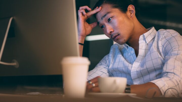 Person in a checkered shirt looking at a computer screen with a focused expression, hand on forehead. The emotional toll of the situation is palpable. Two cups are visible on the desk in the foreground.