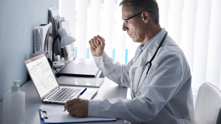 A doctor in a white coat and stethoscope sits at a desk, expertly navigating data management by viewing a laptop and taking notes on a notepad, ensuring the best practices in healthcare are followed.