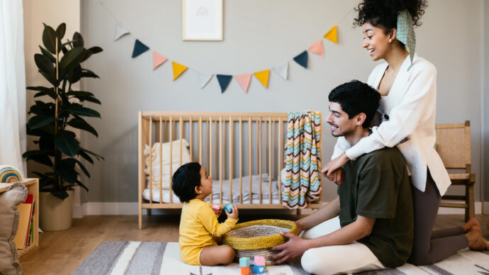 In a cozy nursery room, a family of three enjoys precious moments. The child giggles while playing with toys, beneath a decorative garland on the wall—a serene setting amid the backdrop of declining birthrates.