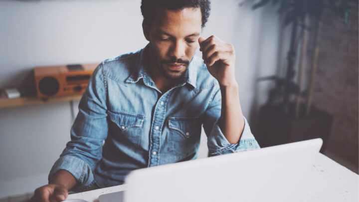 A high-achieving man in a denim shirt sits at a table, focused intently on a laptop screen, his hand resting thoughtfully against his chin. In the background, a plant and an orange stereo add color to the room.