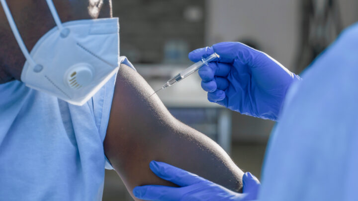 A healthcare worker carefully administers a flu vaccine to a person's arm. Both individuals are wearing protective gloves, with the patient also donning a face mask, adhering to CDC guidelines.