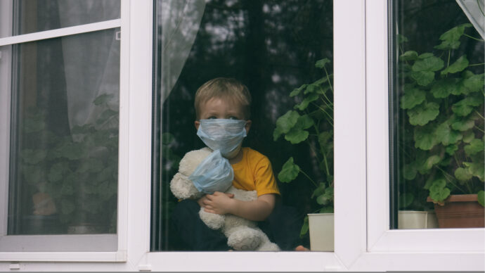 A child wearing a mask sits behind a window, holding a stuffed bear also wearing a mask, surrounded by potted plants, reflecting the cautious spirit of wave 2.