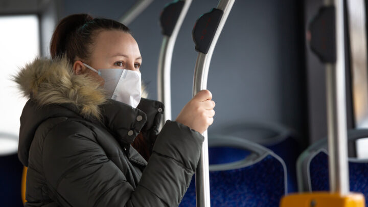 A person wearing a face mask and winter coat is standing on a bus, holding onto a pole, as more people embrace vaccines to stay safe.