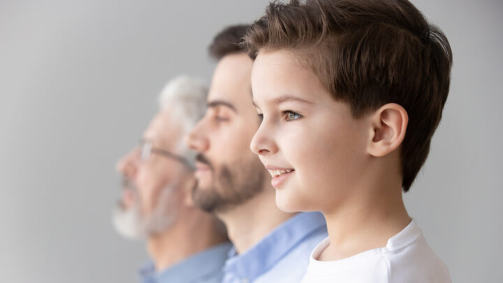A young boy, a middle-aged man, and an older man are lined up in profile, facing left against a blurred gray background, symbolizing the genetic journey of life.