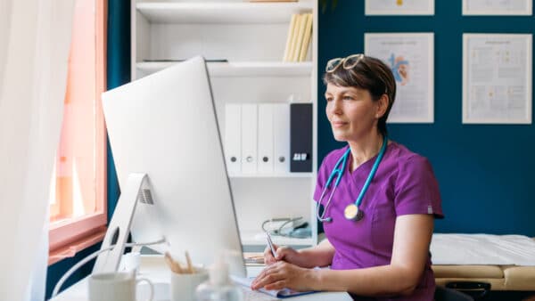 In a well-organized office, a woman in purple scrubs, with a stethoscope around her neck, focuses intently on her computer. Surrounded by shelves and framed certificates, she's leveraging her expertise to debunk myths on HCP social media.