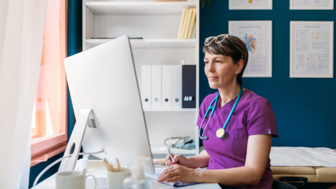 In a well-organized office, a woman in purple scrubs, with a stethoscope around her neck, focuses intently on her computer. Surrounded by shelves and framed certificates, she's leveraging her expertise to debunk myths on HCP social media.