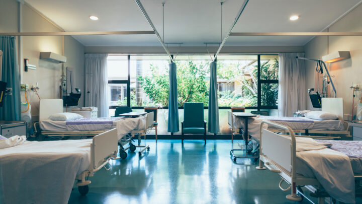 A hospital room with four empty beds, white sheets, and medical equipment stands ready to address health disparities. Large windows in the background reveal greenery outside, reminding us of the world striving for health equity beyond these walls.