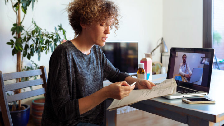 A healthcare professional reads documents at a table with a laptop showing a video call. A phone and a box are also on the table, perhaps part of their 2021 routine.