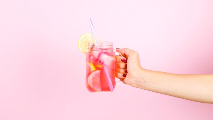 A hand holds a mason jar brimming with refreshing pink lemonade, complete with ice, lemon slices, and a straw against a vibrant pink backdrop—an ideal summer sip to stay healthy.