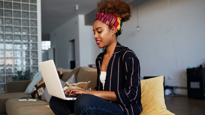 A person sits on a couch, working through the pandemic on a laptop, wearing a striped shirt and a colorful headband. The room has a modern and cozy interior.