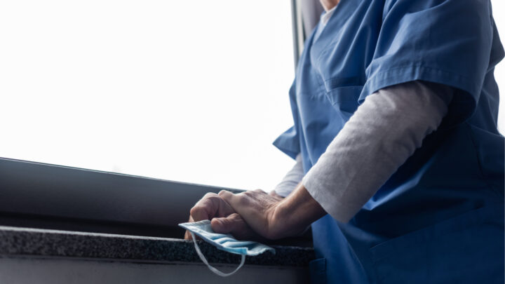 A medical professional in blue scrubs stands by a window holding a surgical mask, embodying the commitment physicians have to patient safety, even amidst the ongoing mask conflict.