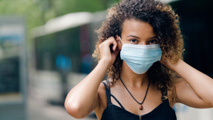 A young person in a black tank top adjusts their surgical mask outdoors, amidst trees and a blurred bus, mindful of the mental health challenges this routine signifies.