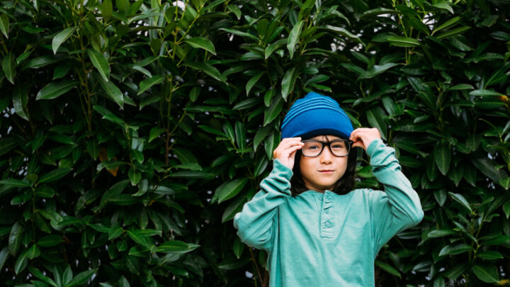 A child with myopia, wearing a green long-sleeve shirt and blue beanie, adjusts large glasses, standing in front of dense green foliage.