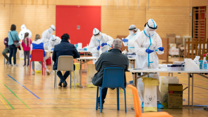 People in protective gear conduct COVID-19 tests at a community testing site, with several patients waiting at tables in a gymnasium setting, highlighting the vital connections between proactive testing and the ongoing vaccine efforts to combat the pandemic.