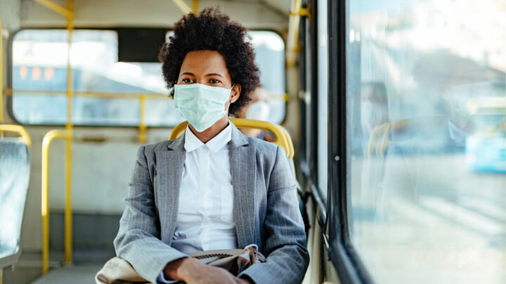 A person of color wearing a face mask and a gray suit sits on a bus, gazing ahead.