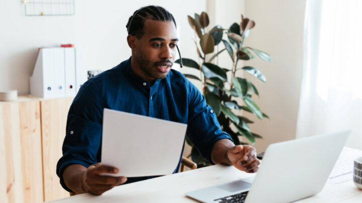 A person with braided hair holds a document while sitting at a desk and looking at a laptop, focused on integrating marketing elements. A plant adds a touch of greenery in the background.