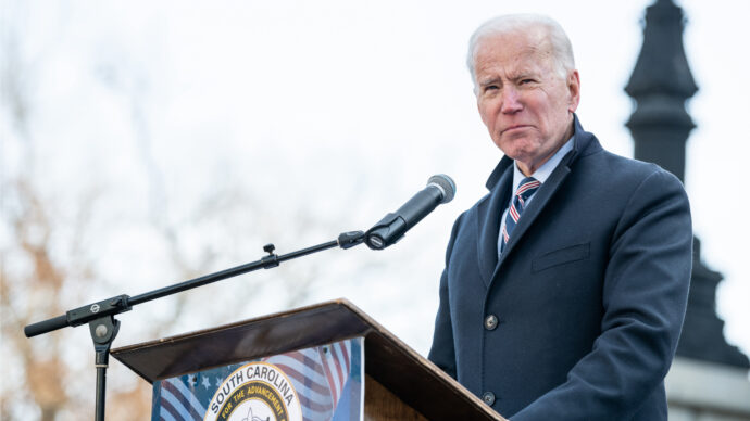 An older man in a suit and tie speaks at a podium outdoors, addressing the impact of Biden's COVID plan on physicians.