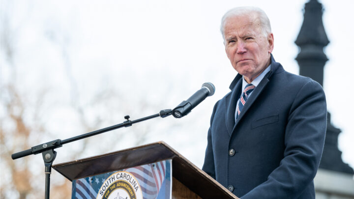 An older man in a suit and tie speaks at a podium outdoors, addressing the impact of Biden's COVID plan on physicians.