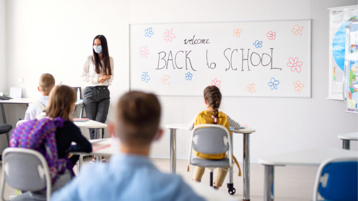 Amid school reopenings, a teacher in a mask stands before a whiteboard with "Welcome Back to School" written on it. Children sit at desks facing the board, as doctors fear another Covid outbreak.