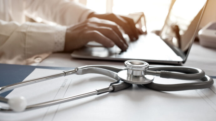 A stethoscope rests on papers, symbolizing health measures, as a doctor types on a laptop in the background.