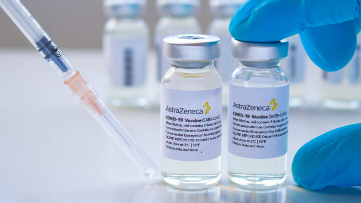 A syringe poised near two AstraZeneca vaccine vials held by a gloved hand, with additional vials blurred in the background. Some physicians express concerns as they oppose the vaccine's widespread use.
