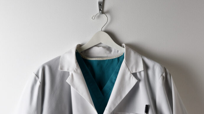 A white lab coat, often seen in a doctor-led primary care setting, hangs on a hanger against a white wall, with a teal scrub shirt underneath.