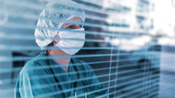 A healthcare worker, likely one of the dedicated physicians, wearing a surgical mask, hair cap, and scrubs peers through a window with blinds, embodying resilience amid COVID stress.