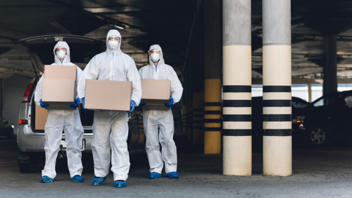 Three individuals in protective suits and masks carry cardboard boxes from a vehicle in a parking garage, illustrating the urgency of the COVID vaccine rollout as some countries remain behind target.