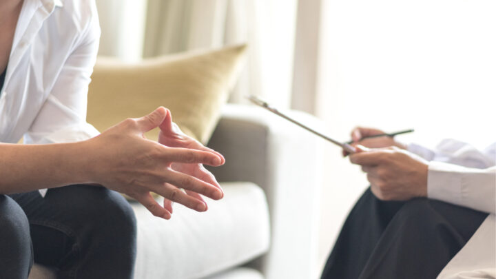 Two people are engaged in a conversation. One has clasped hands, and the other holds a clipboard and pen, offering mental health support while seated comfortably on a sofa with a pillow in the background.