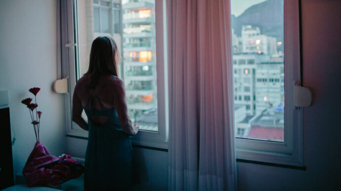 In a dimly lit room, a woman stands gazing out the window at the cityscape during dusk, reflecting on her journey as a doctor working tirelessly to heal patients post-COVID PTSD.