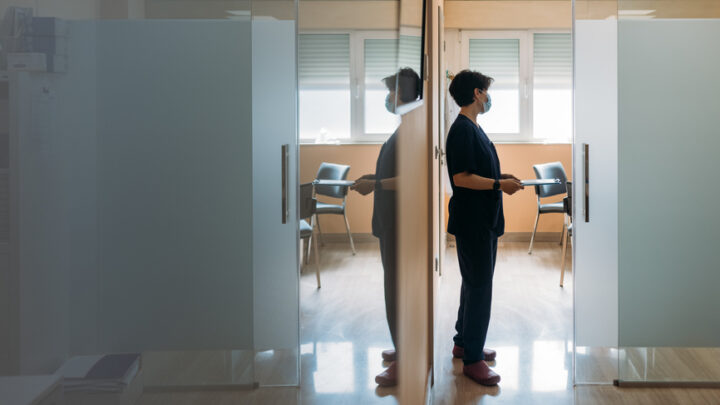 A person in scrubs and mask stands in a hallway, leaning against glass doors, reflecting on the latest clinical trials. Two chairs are visible in a room beyond.