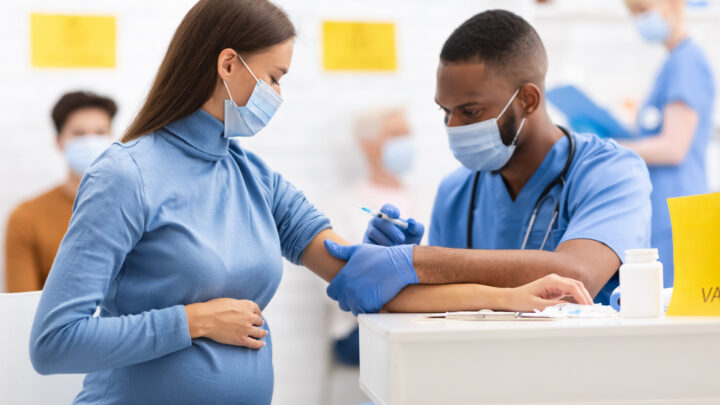 A pregnant woman receives a COVID vaccine in her arm from a healthcare worker wearing blue scrubs and gloves. Both are wearing masks, ensuring the process is safe for pregnancy.