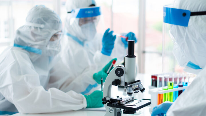 In a lab, scientists in protective gear conduct experiments, with a focus on a microscope in the foreground, as they explore COVID-19 vaccine effectiveness.