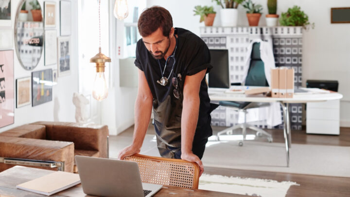 A man stands leaning over a table, looking at a laptop in a bright, modern office with plants and eclectic decor, assessing the value of social media engagement.