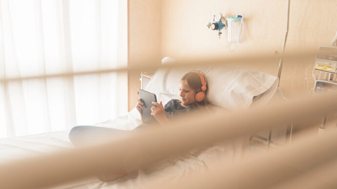 In honor of National Hospital Week, a child with headphones lies comfortably in a hospital bed, absorbed in the world of a tablet.